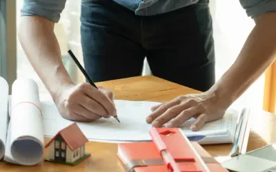 Architekt pisze ołówkiem na planach, obok makieta domu i zwój dokumentów.